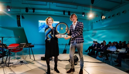 Martina Huijsmans, portfeuillehouder Duurzame Mobiliteit bij de MRDH ontvangt van directeur CROW, Pieter Litjens het gouden wiel. De prijs. Ze staan op een podium en houden de prijs beiden vast.