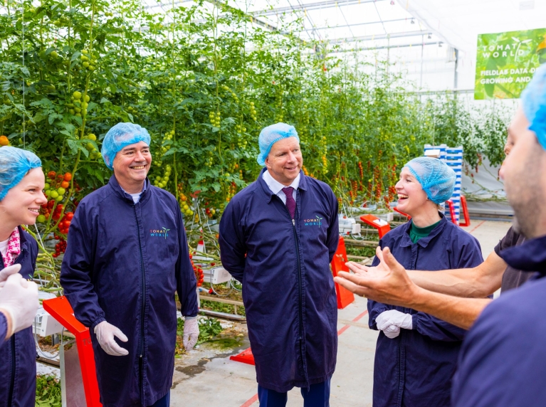 Gedeputeerde Meindert Stolk, wethouder Pieter Varekamp (Westland) en wethouder Maaike Zwart (Delft) zijn in gesprek met medewerkers van TomatoWorld. Ze staan in een kas met beschermende kleding aan.