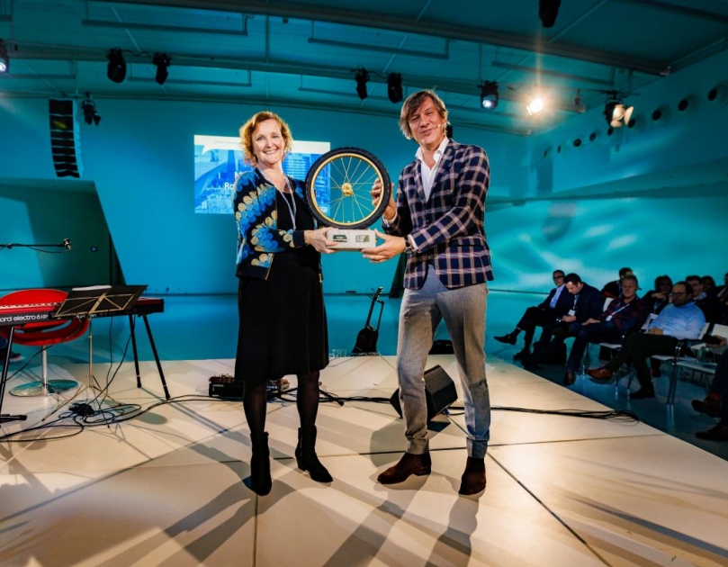 Martina Huijsmans, portfeuillehouder Duurzame Mobiliteit bij de MRDH ontvangt van directeur CROW, Pieter Litjens het gouden wiel. De prijs. Ze staan op een podium en houden de prijs beiden vast.