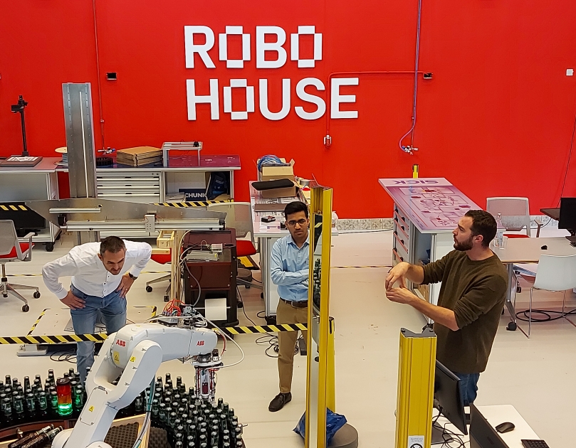 Medewerkers van Robohouse Delft kijken naar een robot die bierflesjes die gevallen flesjes van de lopende band kan oprapen.