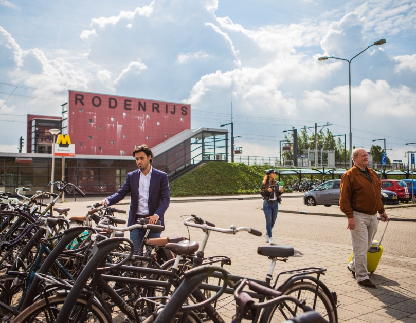 Een man zet zijn fiets in de fietsenstalling bij OV-knooppunt Rodenrijs. Een oudere man rijdt met een rolkoffer richting geparkeerde auto's. Op de achtergrond het metrostation en het parkeerterrein.